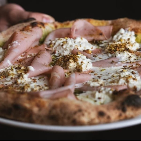A hand holding a freshly baked pizza topped with mortadella, burrata, pesto, and pistachios.