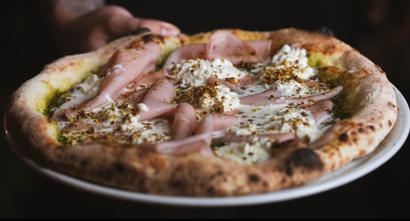A hand holding a freshly baked pizza topped with mortadella, burrata, pesto, and pistachios.