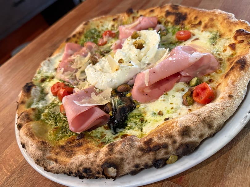 Neapolitan pizza with pesto, mozzarella, prosciutto, cherry tomatoes, and pistachios on a white plate.