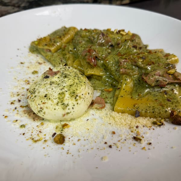 Pasta with green pesto sauce, prosciutto, pistachios, and a burrata ball on a white plate.