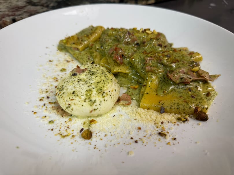 Pasta with green pesto sauce, prosciutto, pistachios, and a burrata ball on a white plate.