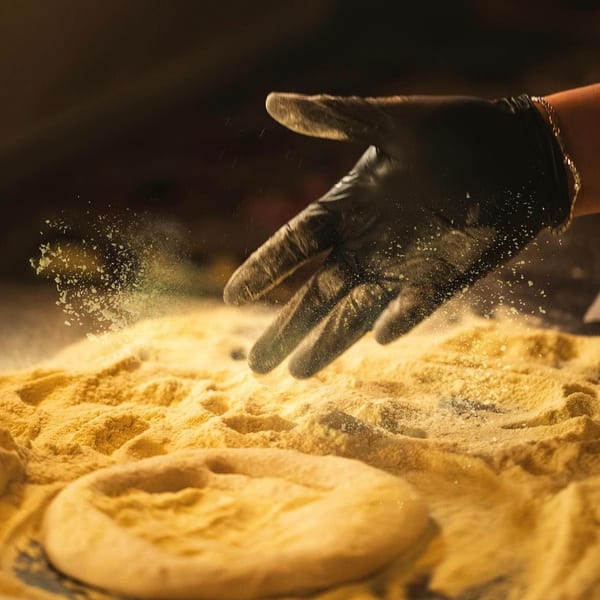 A gloved hand sprinkles flour over a pizza dough base on a floured surface.