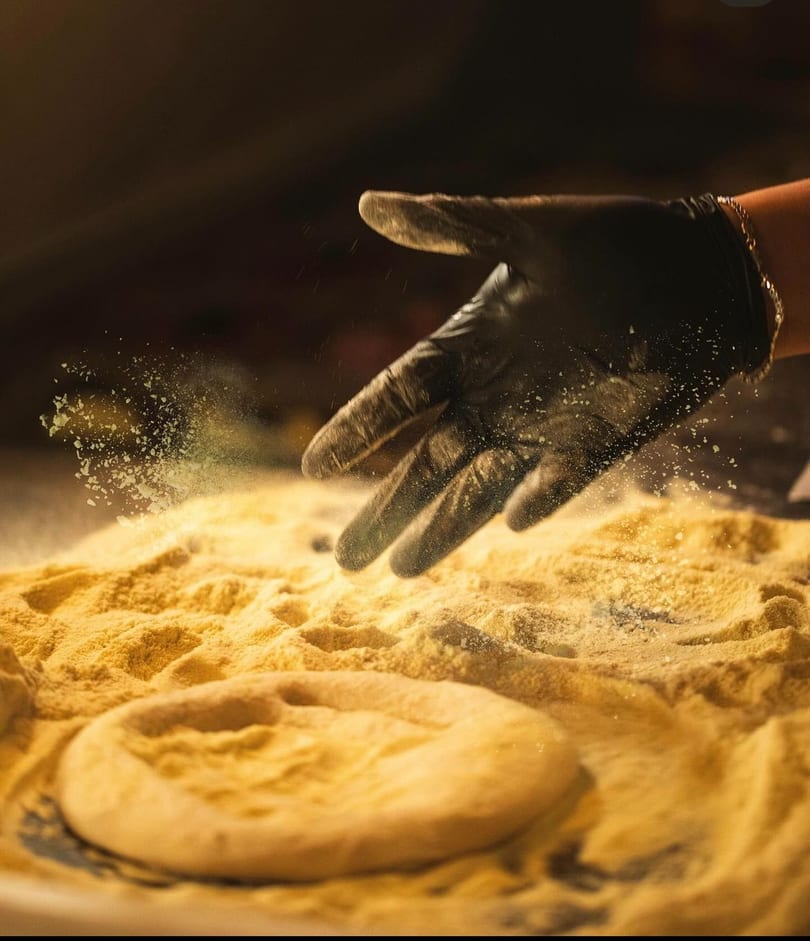 A gloved hand sprinkles flour over a pizza dough base on a floured surface.