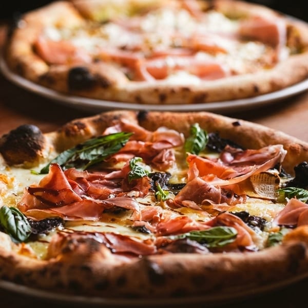Two delicious pizzas with prosciutto, basil, and cheese on a rustic wooden table.