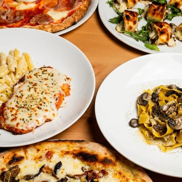 Table spread with Italian dishes: pizzas, chicken parmesan with penne, mushroom pasta, and rolled appetizers.