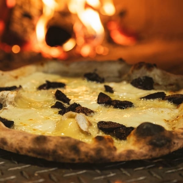 A freshly baked pizza with white cheese and dark toppings sits in a fiery wood-fired oven.
