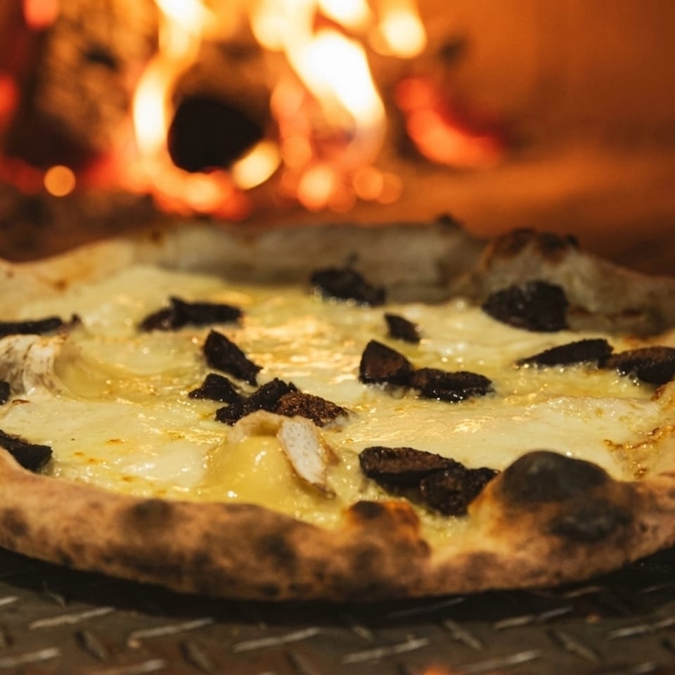 A freshly baked pizza with white cheese and dark toppings sits in a fiery wood-fired oven.