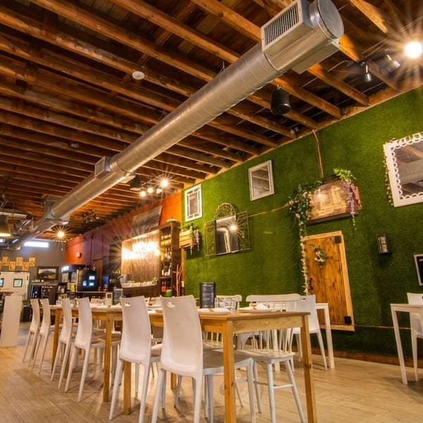 Restaurant interior with wooden beam ceiling, green moss wall, white chairs, and light wood tables.