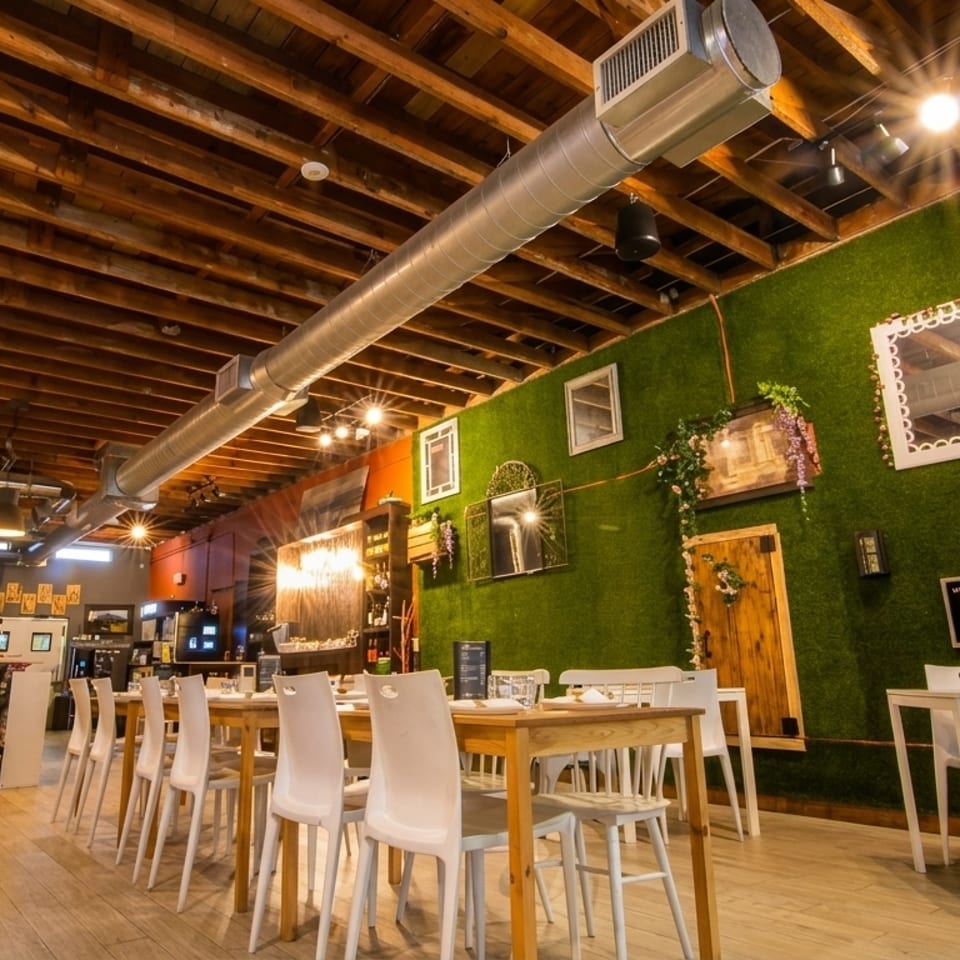 Restaurant interior with wooden beam ceiling, green moss wall, white chairs, and light wood tables.