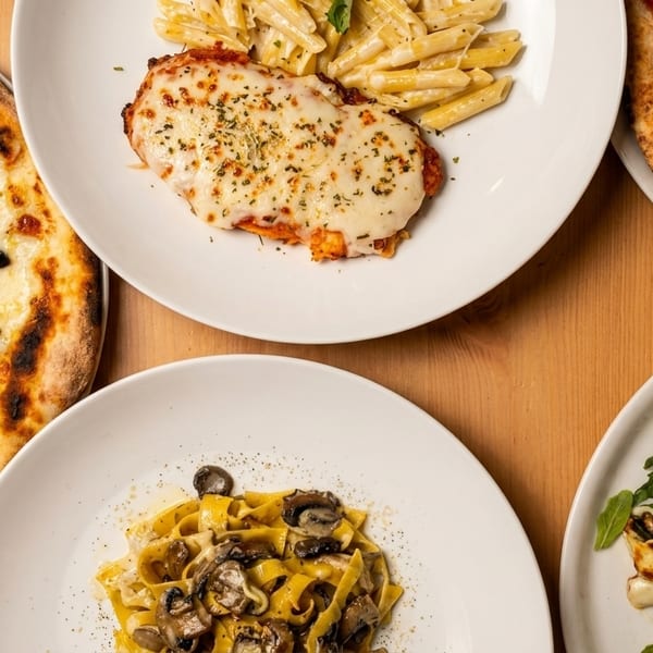 An overhead view of five Italian dishes, including pizzas, pasta, and chicken parmesan, on a wooden table.